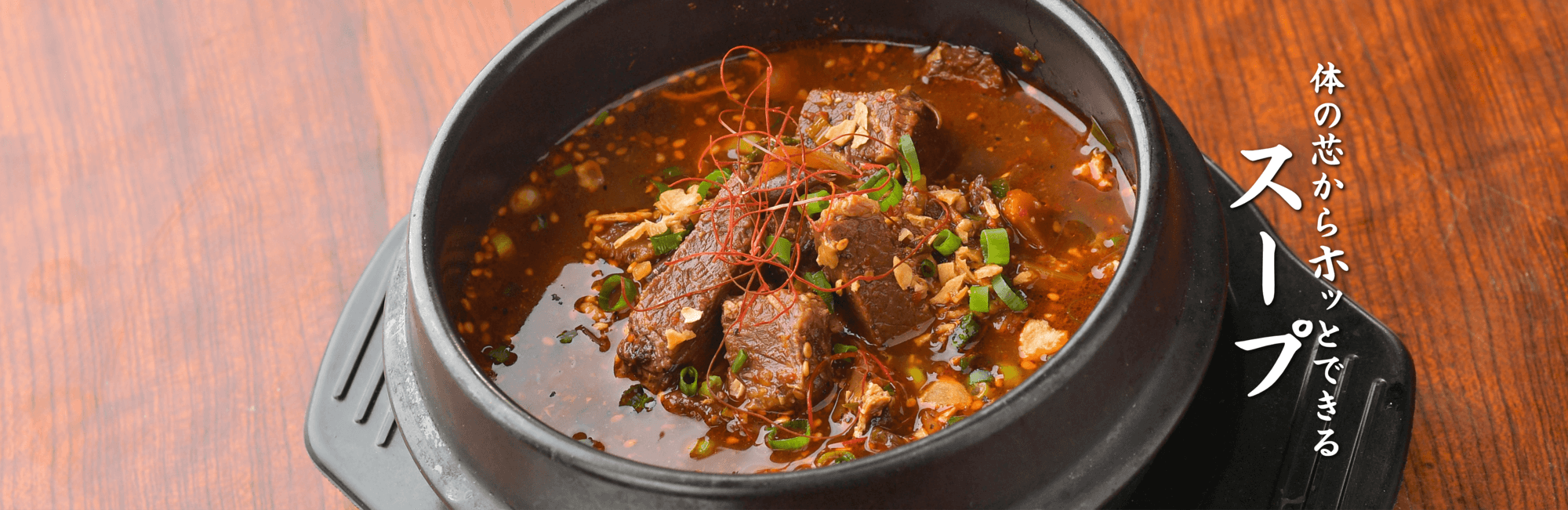 黒い鍋のテグタンスープの写真。真っ赤なスープにたくさんの牛肉とネギ。右側に体の芯からホッとできるスープの文字